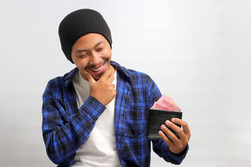 Thoughtful Asian man holds a wallet filled with cash contemplating how to spend it wisely, focusing...