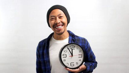 a Cheerful Asian man, dressed in a beanie hat and casual shirt, holds a clock with a broad smile,...