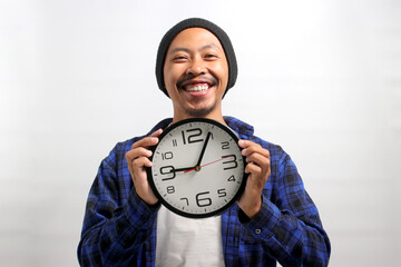 a Cheerful Asian man, dressed in a beanie hat and casual shirt, holds a clock with a broad smile,...