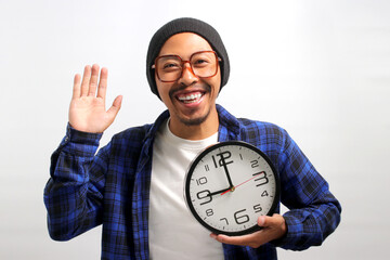 a Cheerful Asian man, dressed in a beanie hat and casual shirt, holds a clock with a broad smile,...