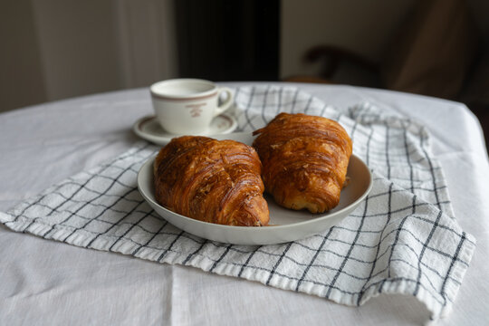 French croissants in a woven basket, complemented by a cup of coffee with milk. A delightful breakfast moment, a blend of pastry perfection.