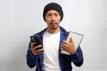 Asian man, dressed in a beanie hat and casual shirt, holds a phone and a book while looking at the...