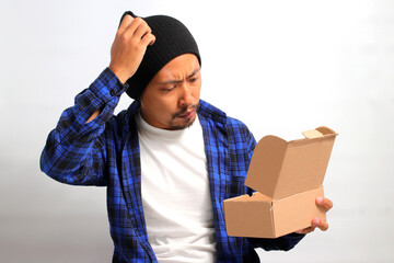 An Asian man, wearing a beanie hat and casual shirt, looks disappointed as he unboxes his delivery,...