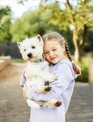 A girl with pigtails holds a small white dog in her arms and smiles happily.