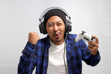 An enthusiastic Asian gamer, adorned in a beanie hat, casual shirt, and headphones, celebrates...