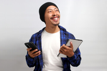 An enthusiastic Asian man, dressed in a beanie hat and casual shirt, holds a phone and a notebook,...
