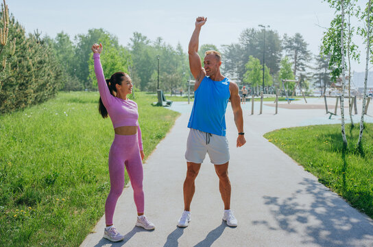A man and woman celebrate fitness success on a sunlit path, their joyful postures and bright workout attire of blue and pink radiating energy and triumph amidst a vibrant green landscape.