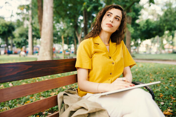 Portrait of pensive young woman sitting on bench in park and drawing sketches in album