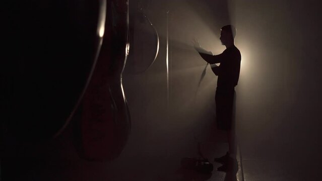 Silhouette of young boxer winds boxing bandages before a match, fight or training. Light in the dark gym.