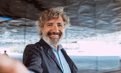 Portrait of a cheerful senior businessman, wears suit, eyeglasses, beard and curly gray hair, broadly smiles and takes a selfie with smartphone outdoors near office building. - Powered by Adobe