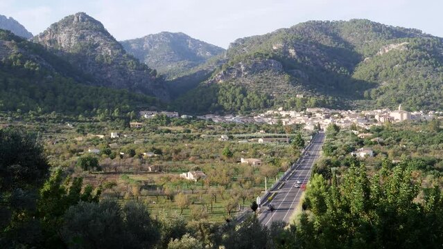 Traveling cars on a serene straight road to picturesque Caimari village, embraced by lush greenery and rolling hills on a beautiful autumn evening in Mallorca. LuPa Creative.