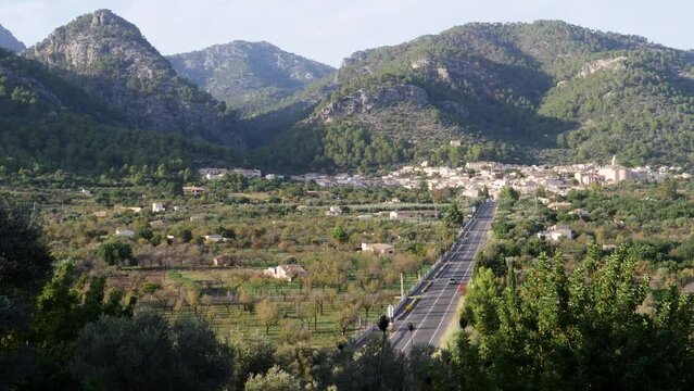 Amidst a serene Mallorcan autumn evening, a single car traverses the picturesque road leading to Caimari, nestled amidst lush hills, capturing the peaceful essence of the village. LuPa Creative.