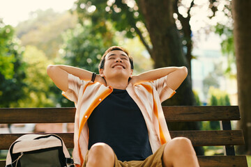 Happy relaxed young man leaning back on park bench and putting hands behind head