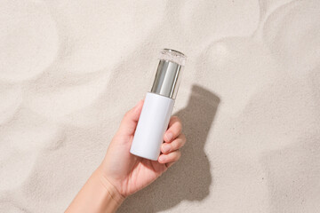 A hand is holding an unlabeled cosmetic bottle on the sandy background. Sophisticated space with a top-down view. Cosmetics advertising.