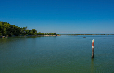 An island in the Grado section of the Marano and Grado Lagoon in Friuli-Venezia Giulia, north east Italy. A channel marker shows the edge of a navigable channel in the shallow waters. August - obrazy, fototapety, plakaty