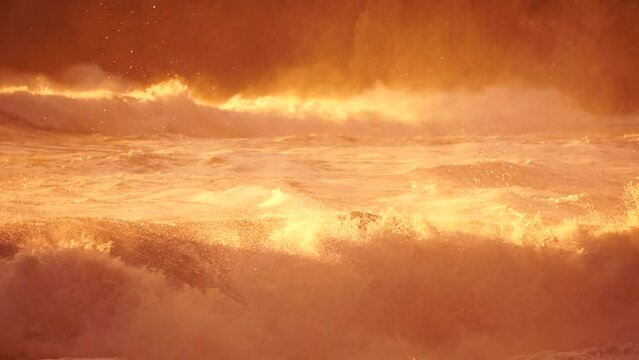 Storm Wave Sunset. Large wave crashing into shore, creating foam and spray at golden burning sunset. Seascape with storm breaking waves. Weather and climate change