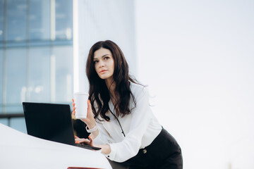 Confident serious young business woman dressed in business style outfit standing outside with laptop, cup of coffee and black classic handbag, female office worker posing against backdrop of cityscape