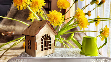 Fototapeta na wymiar miniature toy house in dandelion flowers and watering can. natural background. symbol of family and rural, rustic life. mortgage, construction, rental, property concept. Eco Friendly home. copy space