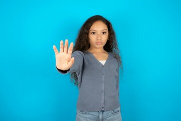 Beautiful teen girl wearing blue jacket over blue background doing stop sing with palm of the hand....
