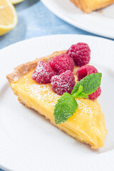 Piece of lemon tart, garnished with fresh raspberry, icing sugar, mint, on plate, vertical closeup