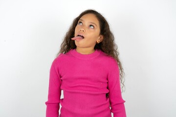Funny Beautiful teen girl wearing pink sweater over white background makes grimace and crosses eyes plays fool has fun alone sticks out tongue. - obrazy, fototapety, plakaty
