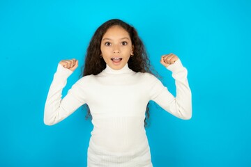 Shocked ecstatic Young beautiful teen girl wearing white turtleneck over blue background win luck...