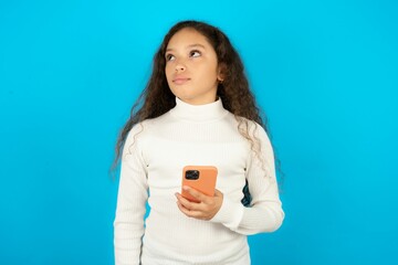 Young beautiful teen girl wearing white turtleneck over blue background holds mobile phone uses high speed internet and social networks has online communication. Modern technologies concept