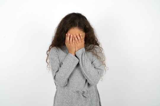 Beautiful teen girl wearing  grey dress over white background covering her face with her hands, being devastated and crying. Sad concept