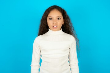 Portrait of dissatisfied Young beautiful teen girl wearing white turtleneck over blue background smirks face, purses lips and looks with annoyance at camera, discontent hearing something unpleasant - obrazy, fototapety, plakaty