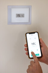 Person holding a smartphone in their hands to adjust their connected thermostat in a French house - 695295813
