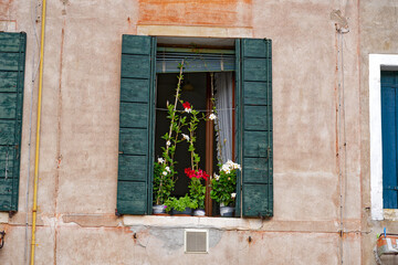 Fototapeta na wymiar Old town of City of Venice with close-up of window with wooden shutters and beautiful flower decoration on a blue cloudy summer day. Photo taken August 6th, 2023, Venice, Italy.
