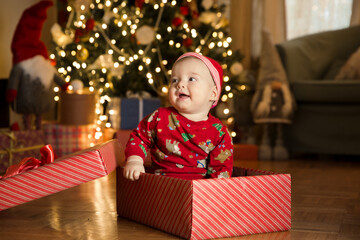 Obraz na płótnie Canvas Happy baby boy in a red cap sitting in a gift box on the background of a Christmas tree on Christmas Eve. Happy Holidays, New Year. Cozy warm winter evening at home. Xmas time