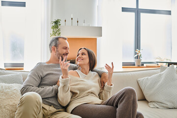 Fototapeta na wymiar cheerful wife talking to smiling husband on cozy couch in living room, modern child-free lifestyle