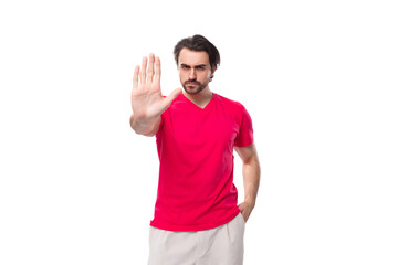 young serious european man dressed in a red corporate t-shirt and white trousers shows a stop gesture with his hand