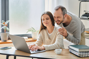 happy man hugging shoulders of smiling  wife working on laptop at home, child-free lifestyle