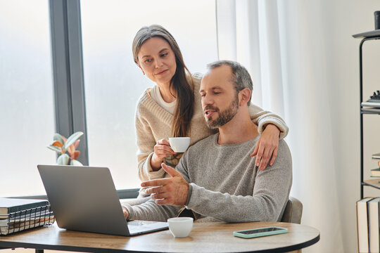 caring wife with coffee cup supporting husband working on laptop in home office, child-free couple