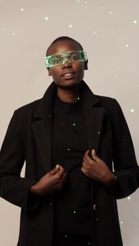 Confident happy young bold African American woman in smart futuristic VR glasses and long black trench coat standing looking at camera against neon light illuminated background