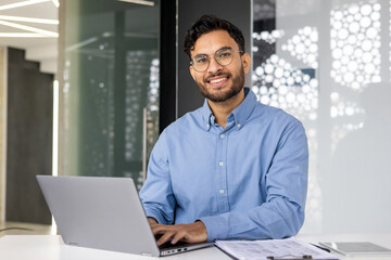 Portrait of young successful businessman inside office, satisfied joyful Indian man looking at...