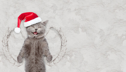 Happy cat wearing red santa hat making snow angel while lying on snow. Empty space for text