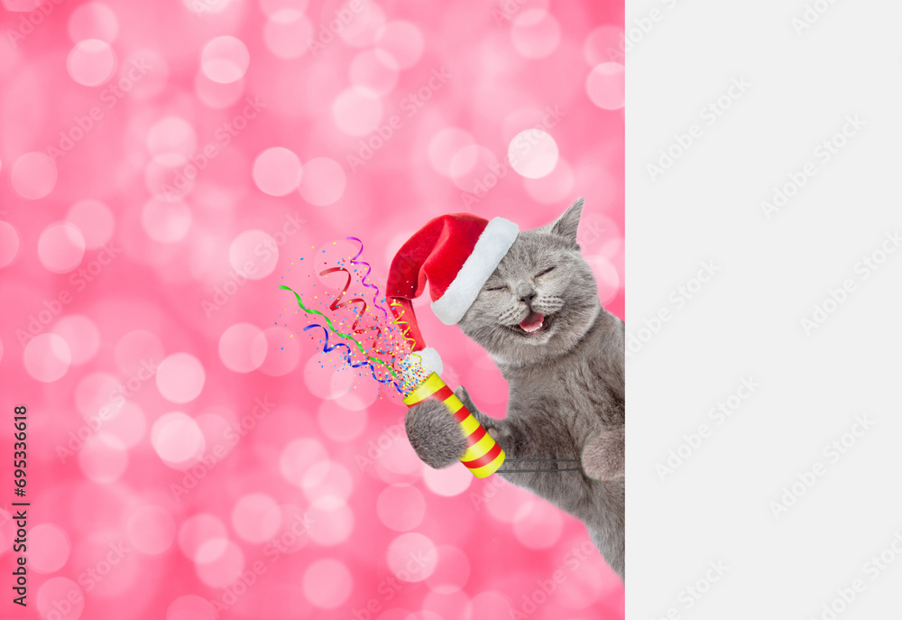Aufkleber Smiling kitten wearing red santa hat holds exploding firecracker and looks from behind empty white banner on festive blurred background