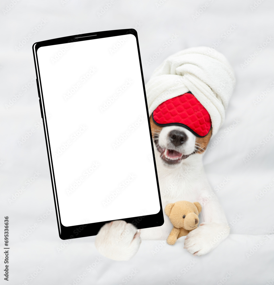 Poster cozy jack russell terrier puppy with towel on it head and with sleeping mask lying on a bed at home 