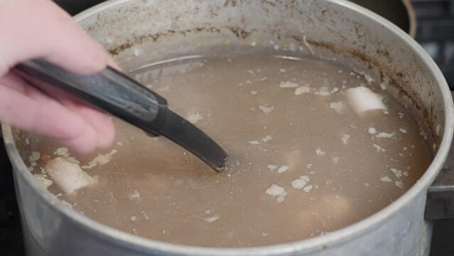 Messy unappetizing bone brooth in aluminum pan, scooping up and stiring in slow motion