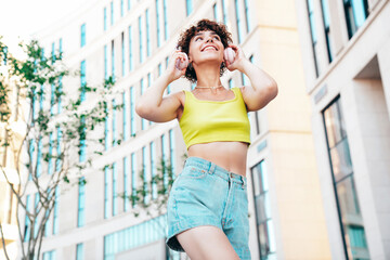 Young beautiful smiling hipster woman in trendy summer clothes. Carefree woman with curls hairstyle, posing in the street at sunny day. Positive model outdoors. Listens music at her headphones