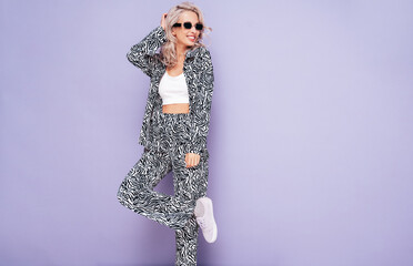 Young beautiful smiling female in trendy summer zebra print costume clothes. Carefree blond woman posing near violet wall in studio. Positive model having fun. Cheerful and happy. In sunglasses
