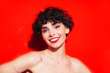 Fashion beauty portrait of young smiling brunette woman with evening stylish  makeup and perfect clean skin. Sexy model with curly short hair posing in studio. With red bright lips. Isolated on red