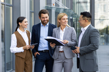 Interracial team of young businessmen and businesswomen standing outside an office center talking to each other, discussing documents and work.