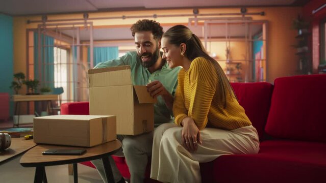 Portrait of Man and Woman Receiving Long-awaited Parcel at Home. Excited Couple Open Cardboard Box while Sitting on a Couch in their Bright Stylish Living Room. Delivery Service for Book Online Store