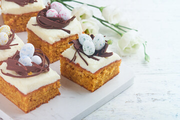 Easter carrot cake bars decorated with chocolate nest and chocolate candy eggs blossoming cherry or apple flowers on rustic light wooden backgrounds. Easter holiday meal. Traditional Easter food.
