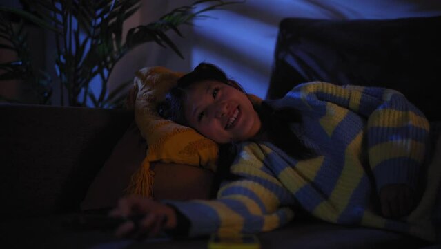 Young Asian woman lying on the sofa quiet watching TV and zapping at home at night. Concentrated female using the television remote control to change channels and watch movies in a dark living room.