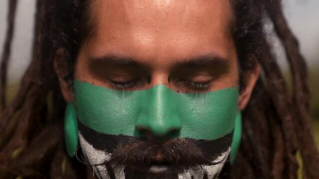 Close up of bearded male warrior with painted face and dreadlocks wearing neck costumes while standing in green park with trees and looking at camera
